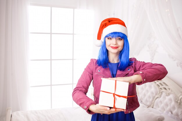 Positieve mooie jonge vrouw pop in de kamer. Rode kerstmuts en kleurrijke kleding. Blauwe pruik Glimlach op het gezicht. Houd de witte doos in handen.