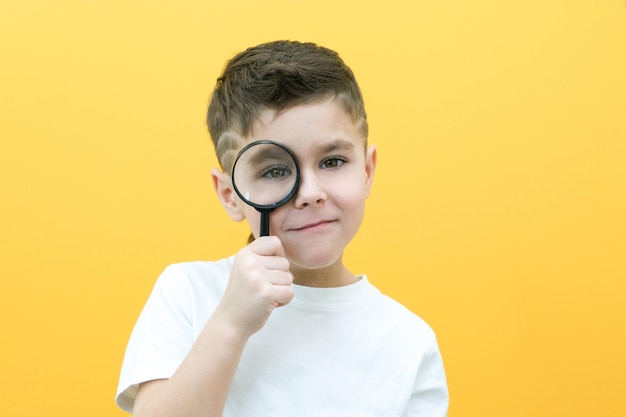 Positieve nieuwsgierige schooljongen in vrijetijdskleding kijkt naar de camera door een vergrootglas op een gele achtergrondverrassing