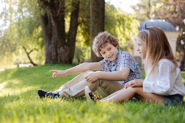 Positieve opgetogen kinderen die op het gras zitten terwijl de jongen naar zijn zus kijkt en iets interessants laat zien