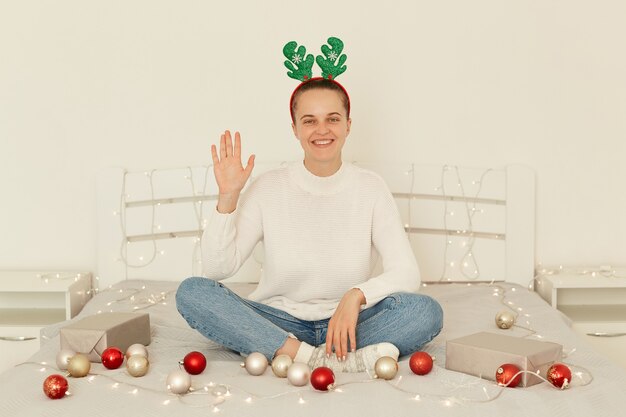 Positieve optimistische vrouw van jonge leeftijd die een witte casual trui en spijkerbroek draagt, met gekruiste benen op bed zit, met de hand zwaait, hallo of dag zegt, camera kijkt met een glimlach.