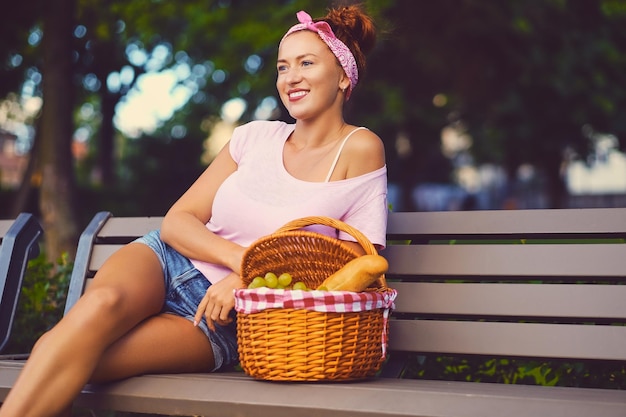 Positieve roodharige vrouw zit op een bankje met een picknickmand vol fruit, brood en wijn.