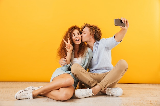 Positieve schattige mensen man en vrouw samen op de vloer zitten en vredesteken tonen terwijl het nemen van selfie op smartphone, geïsoleerd op gele achtergrond