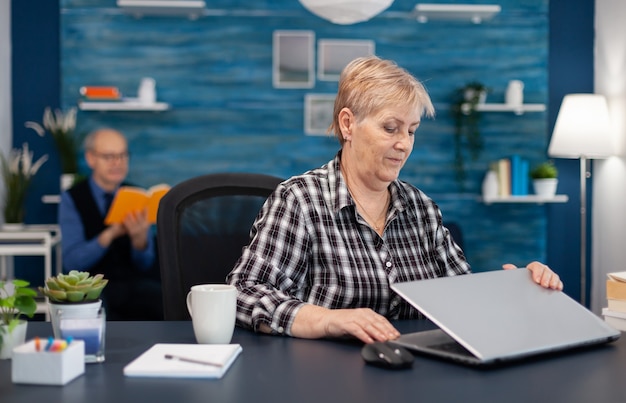 Positieve senior freelancer die laptop opent om thuis te zitten en volwassen man die een boek leest