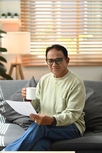 Positieve senior man die papier vasthoudt, betaalt een leningverzekering op een laptop terwijl hij in een gezellige woonkamer zit. ouderen technologieconcept