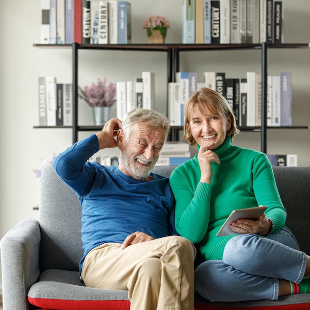 Positieve senior man en volwassen vrouw met tablet glimlachen en kijken naar camera terwijl ze samen op de bank in de woonkamer zitten