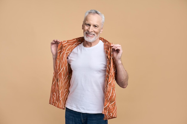 Positieve senior man glimlacht camera kijken houdt handen op oranje shirt geïsoleerde beige achtergrond