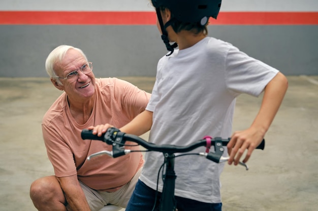 Positieve senior man in vrijetijdskleding en bril met anonieme jongen in helm op fiets samen tijd doorbrengen op parkeerplaats