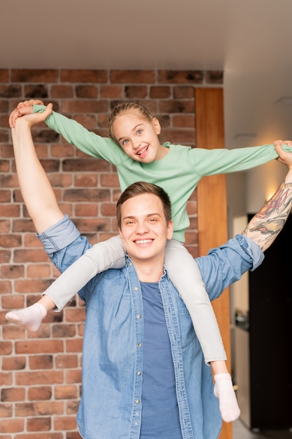 Positieve speelse jonge vader met tatoeage op arm en dochter met gevlochten haar plezier samen thuis, opgewonden meisje zittend op de nek van vader