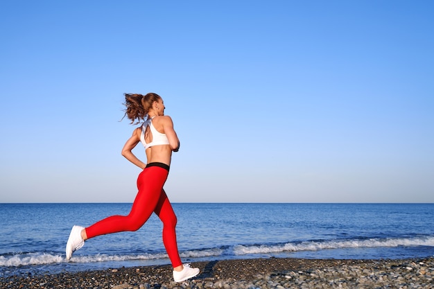 Positieve sportieve vrouw op een zomerochtend joggen op het strand in rode legging op de achtergrond van de zeekust