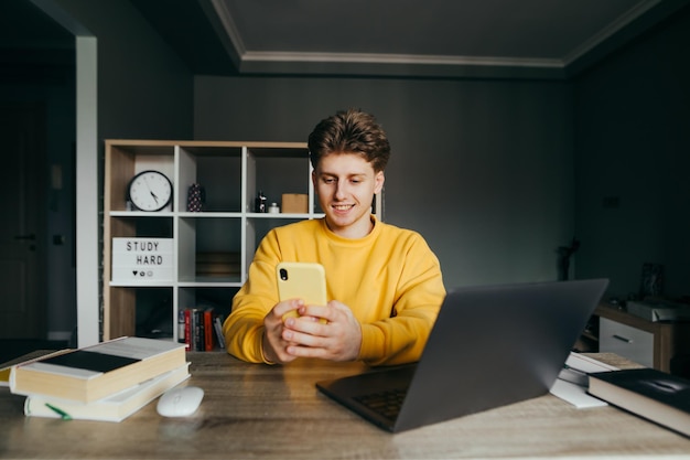Positieve student studeert thuis in een schilderij en gebruikt een smartphone met een glimlach op zijn gezicht zittend aan een tafel in de slaapkamer met een laptop en boeken Online training in quarantaine