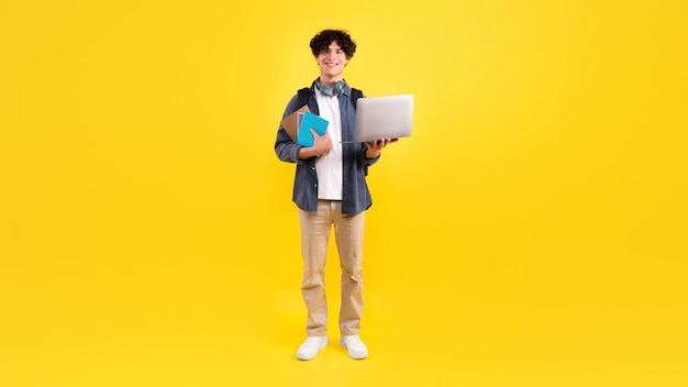 Positieve studentenkerel die laptop en leerboeken over gele achtergrond houdt