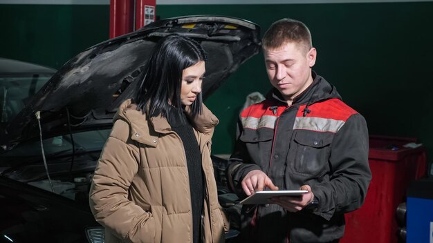 Positieve technicus toont prijslijst op tablet aan vrouwelijke klant in de buurt van kapotte auto met open motorkap bij modern autoreparatieservicestation