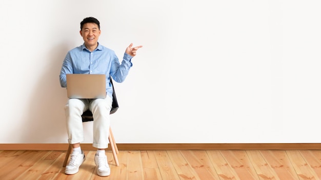 Positieve volwassen aziatische man zittend op een stoel met laptop en wijzend op vrije ruimte terwijl studiomuur volledige lengte