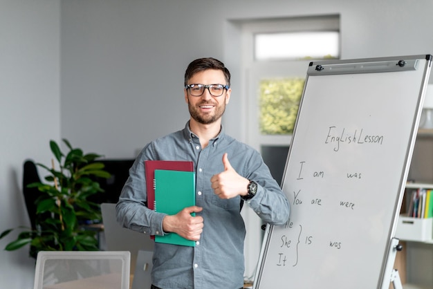 Positieve volwassen mannelijke leraar die duim laat zien terwijl hij in de buurt van schoolbord staat en nieuwe online aanbeveelt