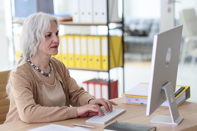 Positieve volwassen werknemer werkt op de computer aan het bureau in het kantoor van het bedrijf, vrouwelijke ondernemer doet dat