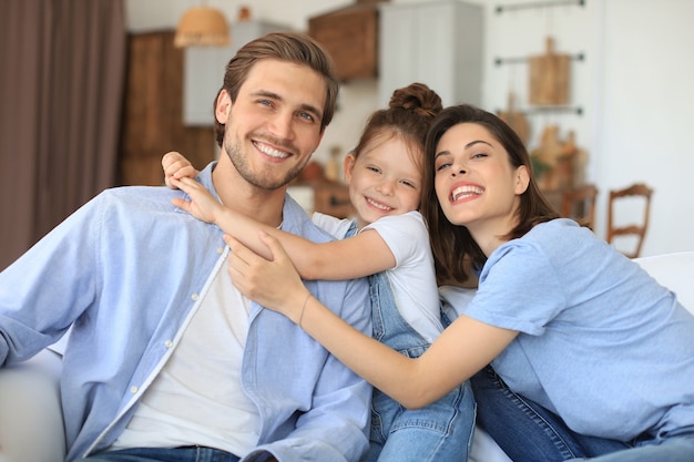 Positieve vriendelijke jonge ouders met een lachend dochtertje dat samen op de bank zit terwijl ze in het weekend thuis ontspannen.