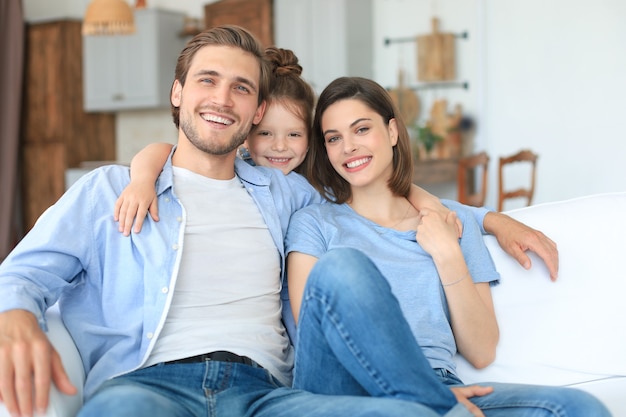 Positieve vriendelijke jonge ouders met een lachend dochtertje dat samen op de bank zit terwijl ze in het weekend thuis ontspannen.