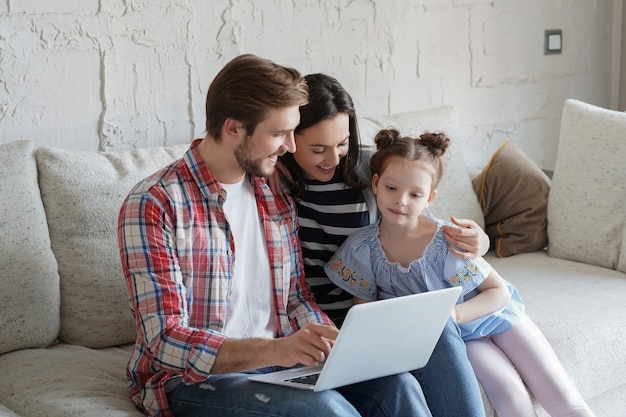 Positieve vriendelijke jonge ouders met lachende dochtertje zittend op de bank samen kijken naar foto's op laptop, thuis ontspannen in het weekend.