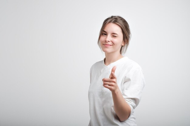 Positieve, vrolijke jonge blanke vrouw die een wit casual T-shirt draagt, met haar ogen knippert en naar de camera glimlacht