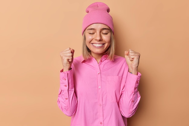 Positieve vrouw balt vuisten en anticipeert op goede resultaten houdt ogen gesloten glimlacht tandjes draagt roze hoed en shirt voelt als winnaar viert geweldig nieuws staat binnen tegen bruine muur