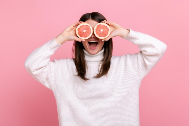 Positieve vrouw die de ogen bedekt met twee halve plakjes grapefruit, detox en gezond rauw vers voedsel, met een witte casual stijltrui. Indoor studio opname geïsoleerd op roze achtergrond.
