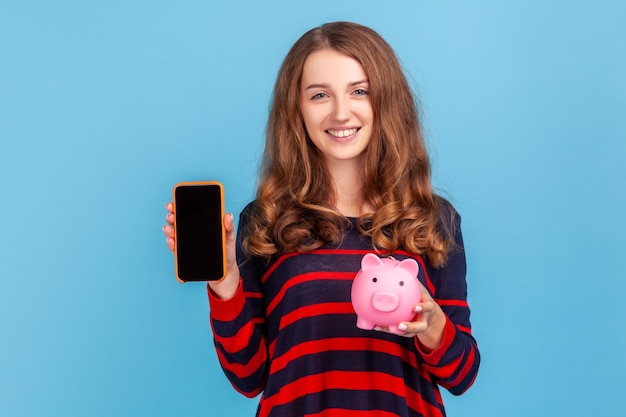 Positieve vrouw draagt gestreepte casual stijl trui met spaarvarken om geld te besparen en smartphone met leeg scherm voor reclame Indoor studio shot geïsoleerd op blauwe achtergrond