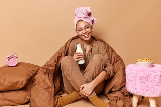 Positieve vrouw drinkt verse melk voor het ontbijt draagt badhanddoek op hoofdpyjama en sokken poseert onder zachte deken op comfortabel bed thuis geniet van goedemorgen geïsoleerd over beige achtergrond