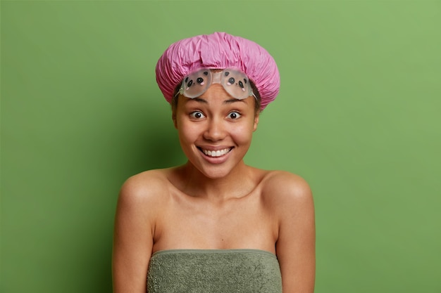 Positieve vrouw glimlacht in grote lijnen gaat douchen draagt badmuts waterdichte bril gewikkeld in handdoek heeft gezonde gladde huid geïsoleerd over levendige groene muur