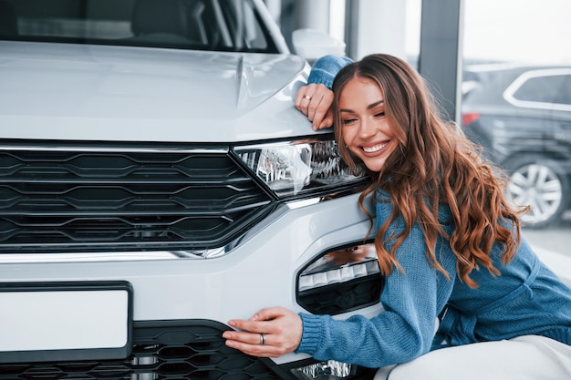 Positieve vrouw in blauw shirt omarmen haar gloednieuwe auto In autosalon