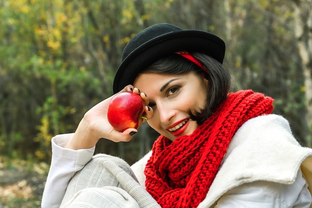 Positieve vrouw in zwarte hoed en rode sjaal met een rode appel in haar hand glimlacht en kijkt naar de camera