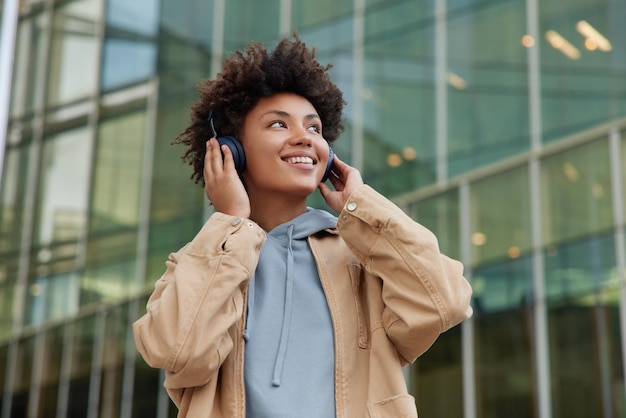 Positieve vrouw met krullend haar geniet van het luisteren naar muziek via een koptelefoon draagt een hoodie en een jas glimlacht gelukkig kijkt ergens opzij poses tegen een modern glazen gebouw geniet van favoriete audiotrack