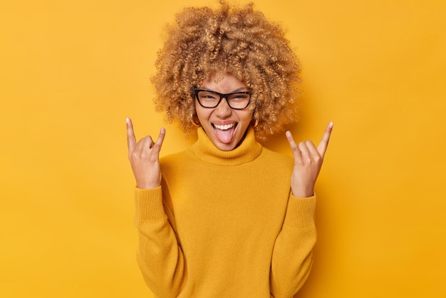 Foto positieve vrouw toont rock n roll gebaar heavy metal teken geniet van iets geweldigs steekt tong geniet van rockmuziek draagt een bril casual jumper geïsoleerd over gele achtergrond heeft plezier.