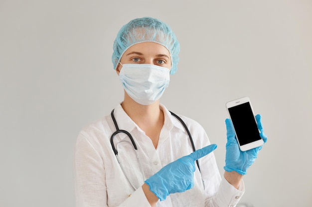 Positieve vrouwelijke arts die medische pet, handschoenen, chirurgisch masker en toga draagt, staand en mobiele telefoon toont met een leeg scherm voor reclame of promotie.