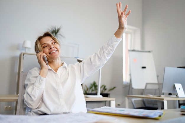 Foto positieve zakenvrouw in wit overhemd praten op de mobiele telefoon en zwaaien met haar hand