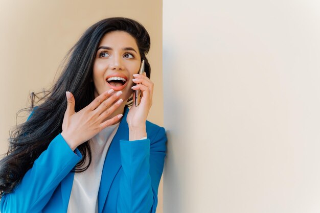 Positieve zakenvrouw opent mond van interesse raakt kin aan kijkt opzij gefascineerd door geruchten heeft telefoongesprek met goede vriend roddels over het laatste nieuws draagt formeel blauw kostuum