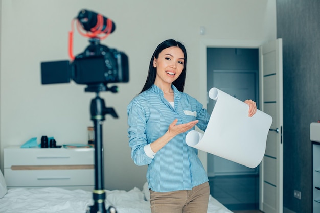 Positieve zelfverzekerde jongedame die staat met een rol architectonisch papier en glimlacht naar de camera op statief terwijl ze het demonstreert