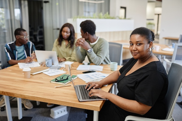 Positieve zwarte vrouwelijke ondernemer die aan laptop werkt wanneer haar collega's een briefingsessie hebben