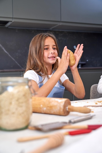 エプロンに長い髪をしたポジティブな愛らしい子供が、テーブルに座り、キッチンでペストリーを準備しながら、笑顔で手で生地をこねる