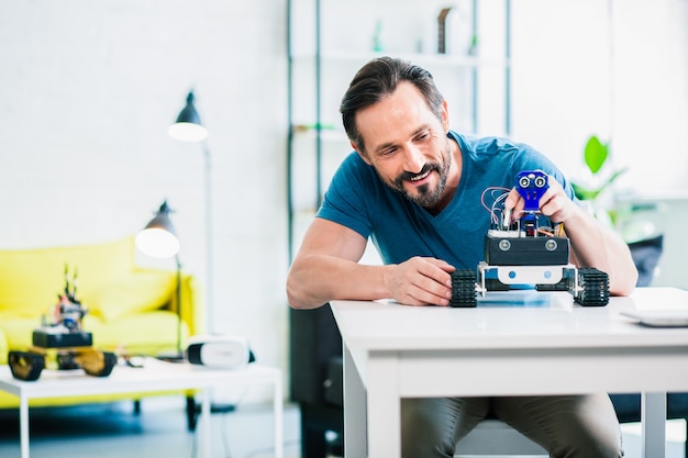 Positive adult bearded man sitting at the table and enjoying robotic engineering