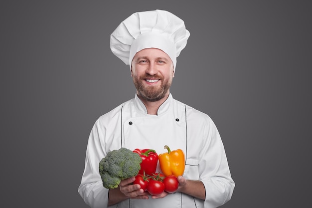 Positive adult bearded professional cook in white chef uniform demonstrating various colorful fresh vegetables for healthy meal preparation against gray background