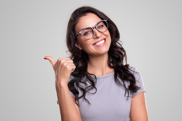 Positive adult brunette in casual dress and trendy eyeglasses smiling happily while pointing aside with thumb up against gray background