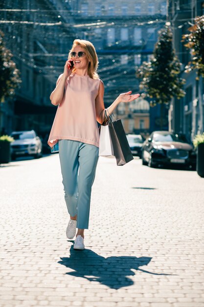 Positive adult lady with shopping bags strolling outdoors and smiling during the phone conversation