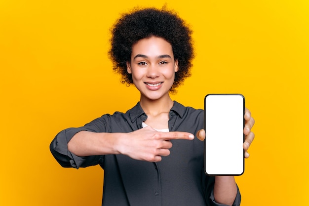 긍정적 인 아프리카계 미국인 또는 브라질 여성은 프레젠테이션 또는 광고를 위해 빈  모형 화면으로 스마트 폰을 들고 고립 된 노란색 배경에 카메라 스탠드에 손가락을 겨누고 미소 짓습니다.