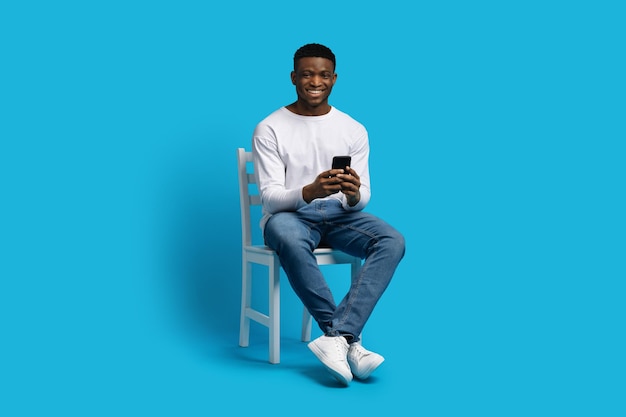 Positive african american guy sitting on chair using smartphone