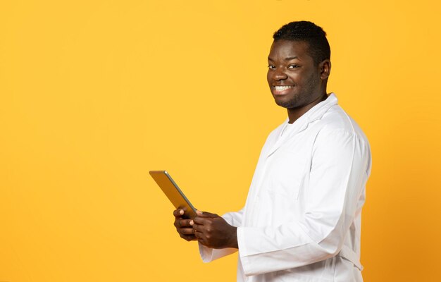 Positive african american man doctor holding digital tablet yellow background