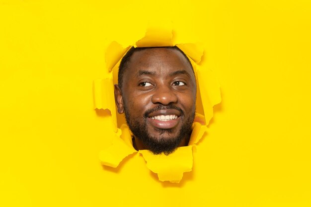 Positive african american man looking aside and smiling while tearing hole in yellow paper background copy space