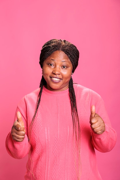 Positive african american model giving approved sign standing over pink background in studio. Happy cheerful woman with joyful facial expression showing thumbs up gesture, person doing like sign