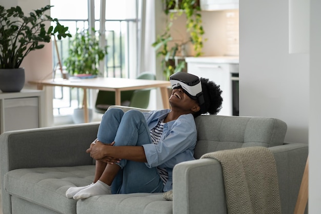 Positive African American woman teenager in futuristic VR helmet laughing and relaxing sits on sofa