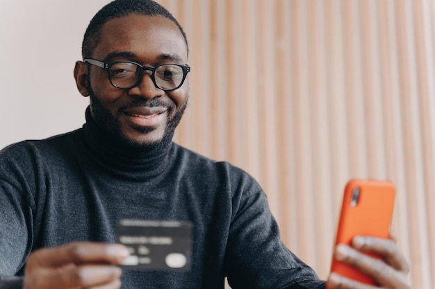 Positive African entrepreneur in glasses paying online with credit card ordering on cellphone smiling millennial guy using mobile banking app for transaction