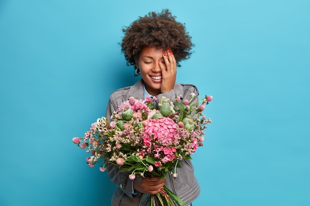 Positive Afro American woman holds beautiful bouquet of flowers received on birthday covers face with hand dressed in grey jacket isolated over blue wall